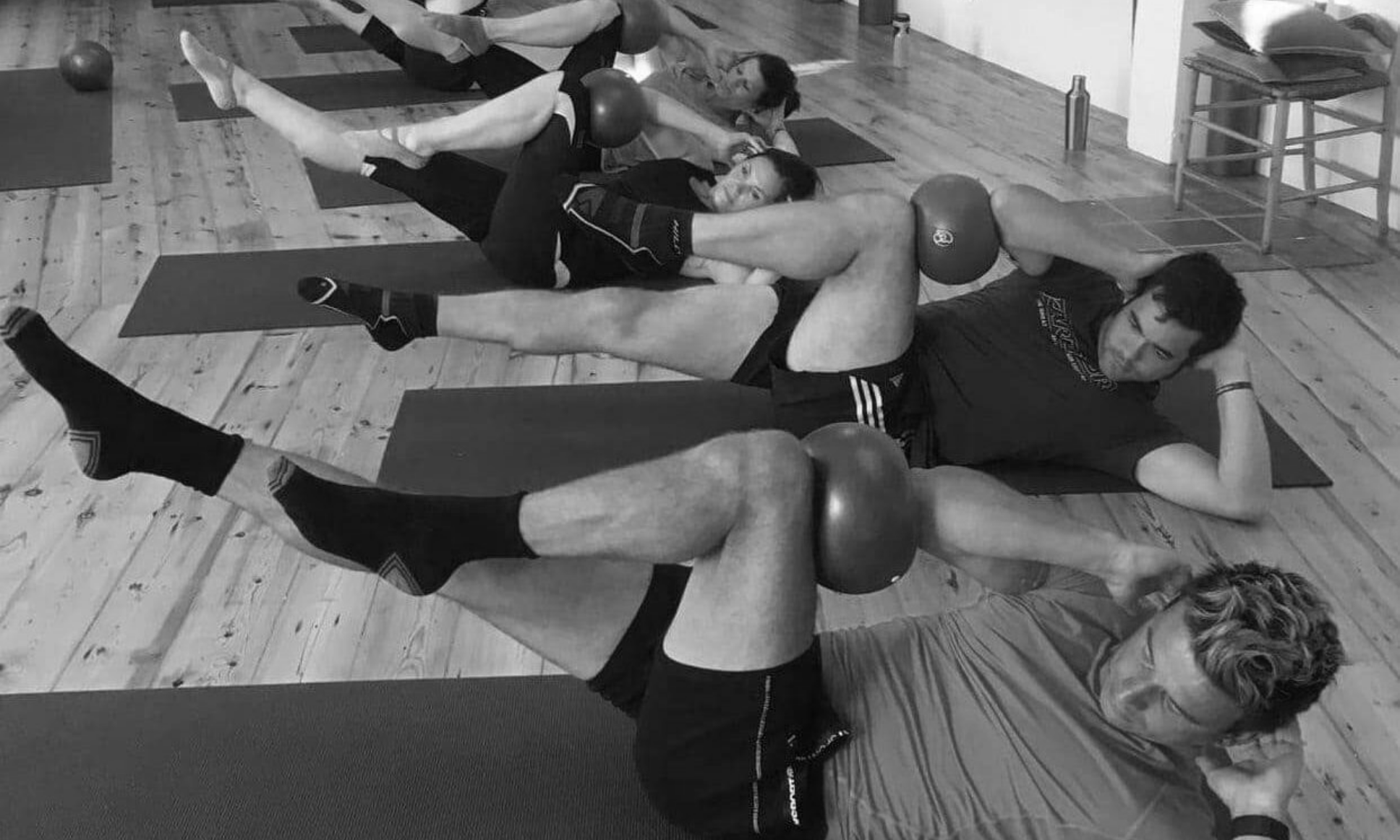 A men's mat class on the studio boards, legs raised with small balls