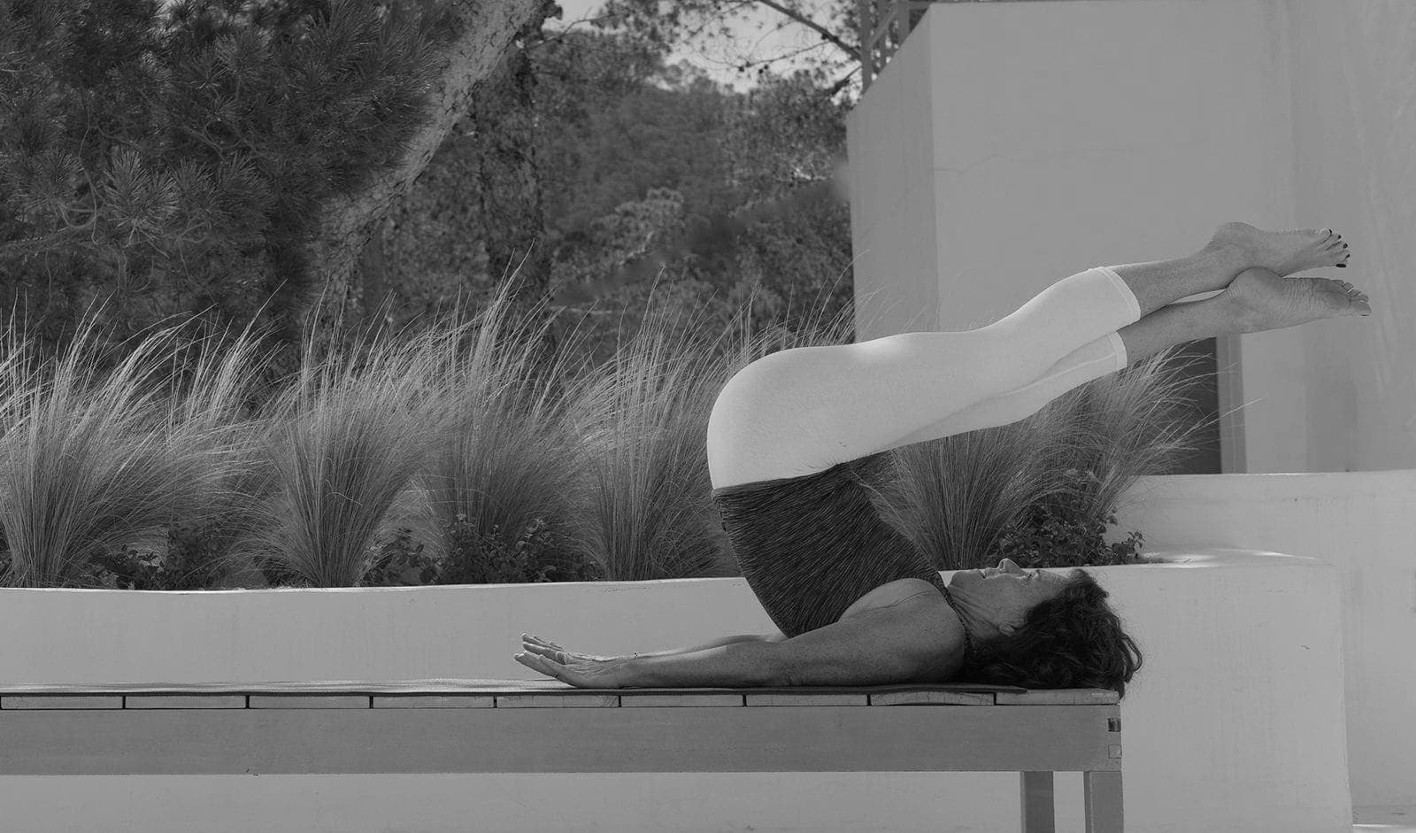 Caroline McGee in a jackknife on a wooden bench, legs extended over ornamental grasses